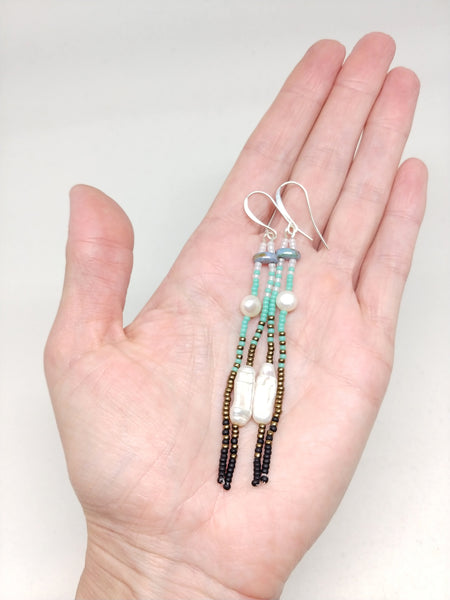 Hand holding Calm Dangles: Silver-Plated ear wires, pearls, and white, blue, bronze and black seed beads.