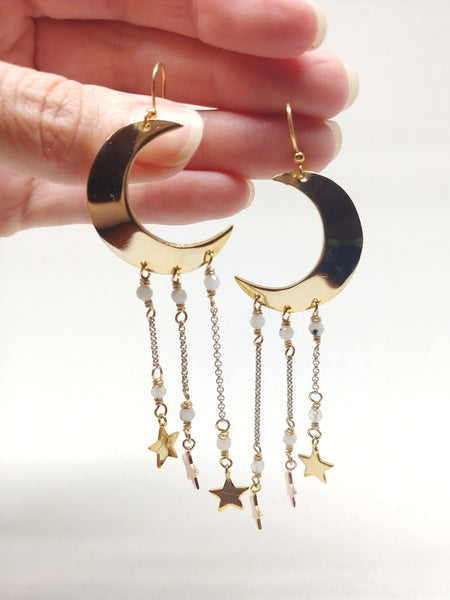 Large gold crescent moon earrings with shiny stars dangling from underneath by silver chain and moonstone accents, being held for scale.